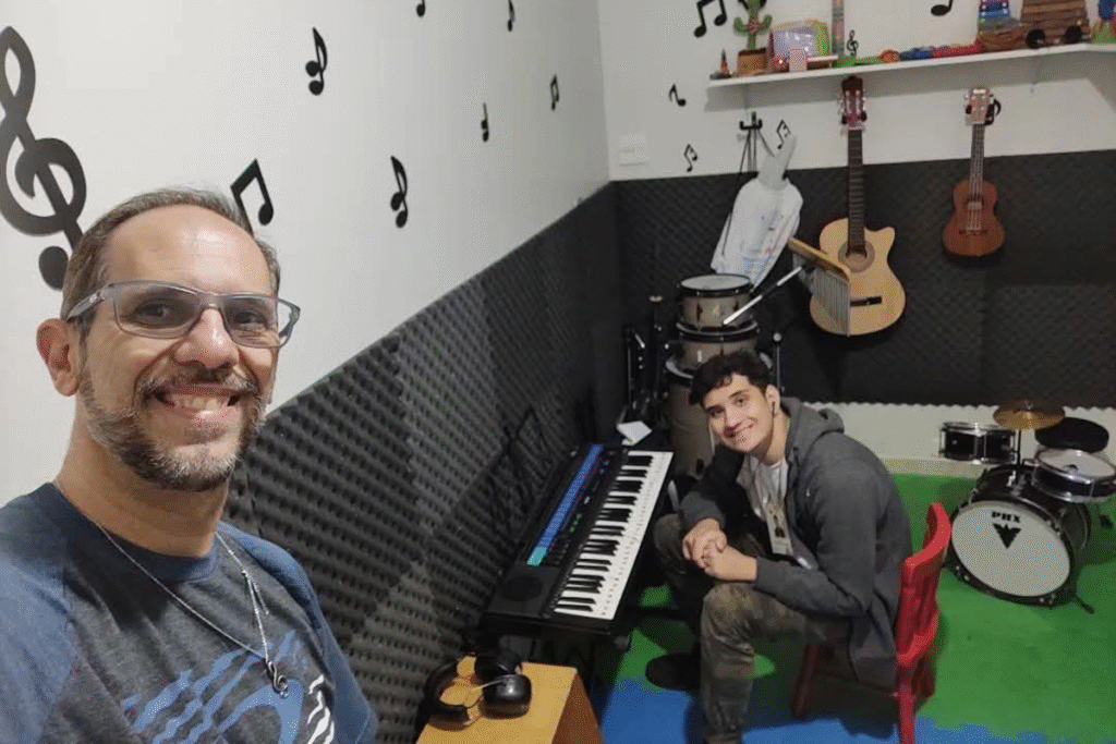 Foto de Marcelo com seu filho Rafael em uma sala cheia de instrumentos musicais. Rafael está sentado em uma cadeira baixa em frente a um teclado. Marcelo está em primeiro plano fazendo a self. Ambos estão sorrindo.