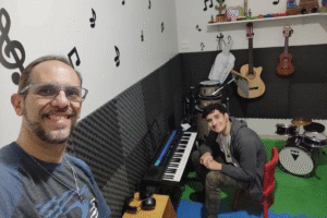 Foto de Marcelo com seu filho Rafael em uma sala cheia de instrumentos musicais. Rafael está sentado em uma cadeira baixa em frente a um teclado. Marcelo está em primeiro plano fazendo a self. Ambos estão sorrindo.