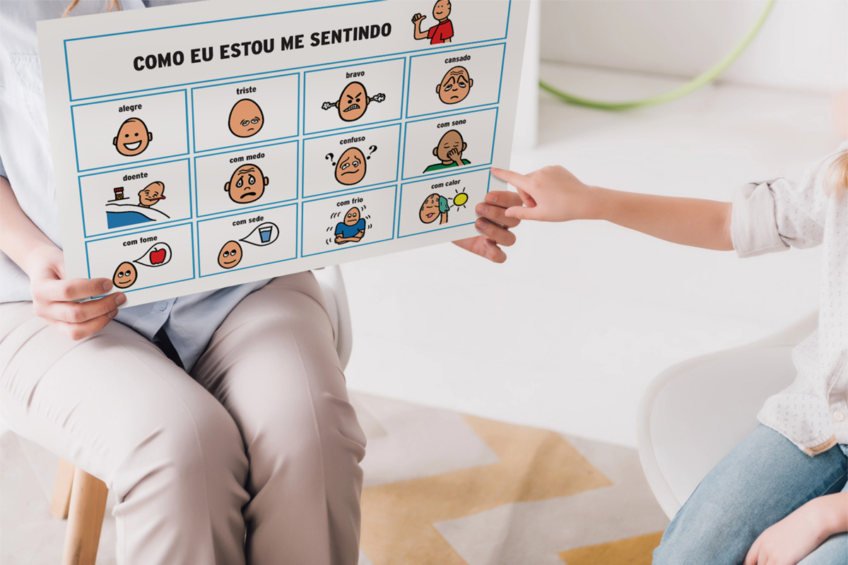 Foto de uma psicologa mostrando uma prancha de comunicação alternativa com os sentimentos a uma criança, e ela está apontando para figura que está com sono.