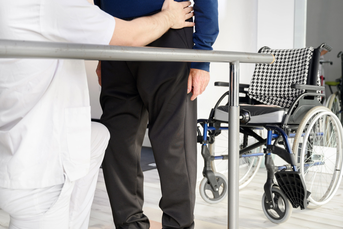 Foto de uma terapeuta ocupacional ajudando seu paciente a andar. Ao fundo vemos sua cadeira de rodas