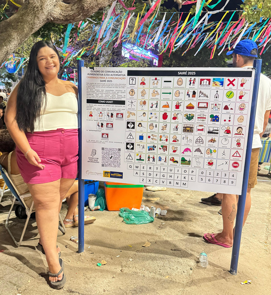 Foto de Glaice ao lado da placa de Comunicação Alternativa do Festival Saire 2025.