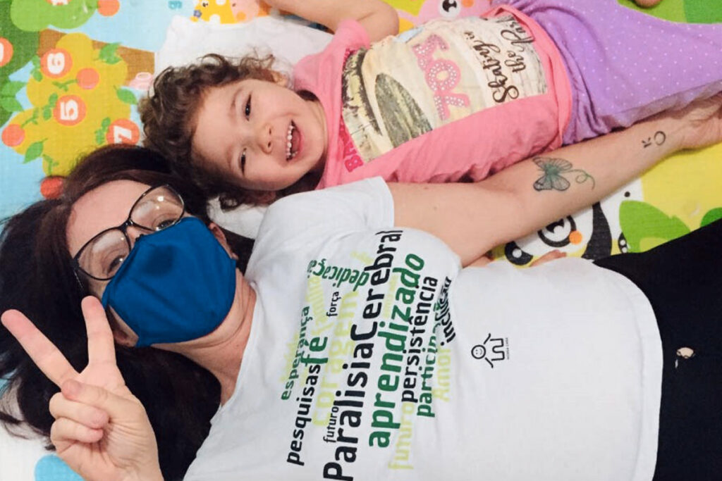 Foto de Cida deitada ao lado de sua aluna Cecilia. Ambas olham para câmera sorrindo. Cida está usando máscara e faz um sinal de V com a mão.