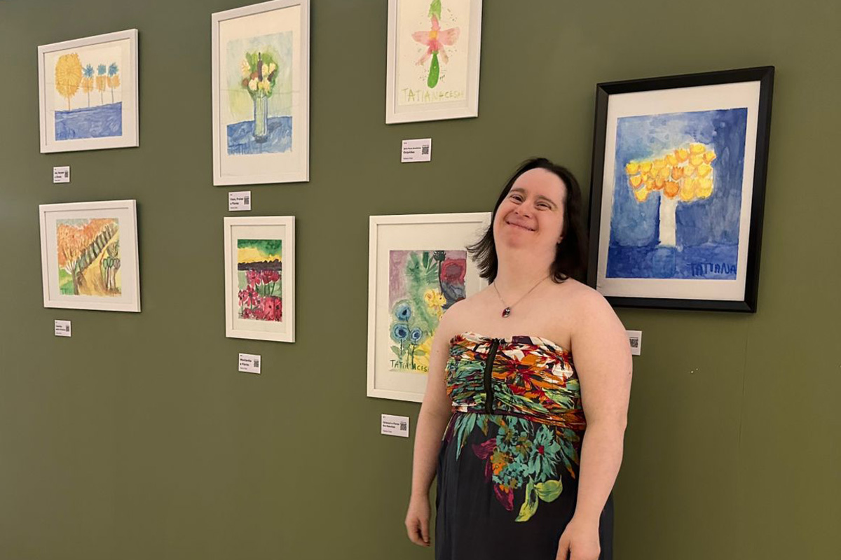 Foto de Tatiana sorrindo em frente a uma parede cheia de aquarelas que ela fez.