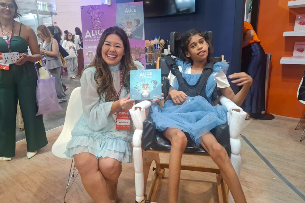 Foto de Valeria segurando o livro Alice no País das Palavras Mágicas ao lado de Maju, que foi quem a inspirou a escreve-lo
