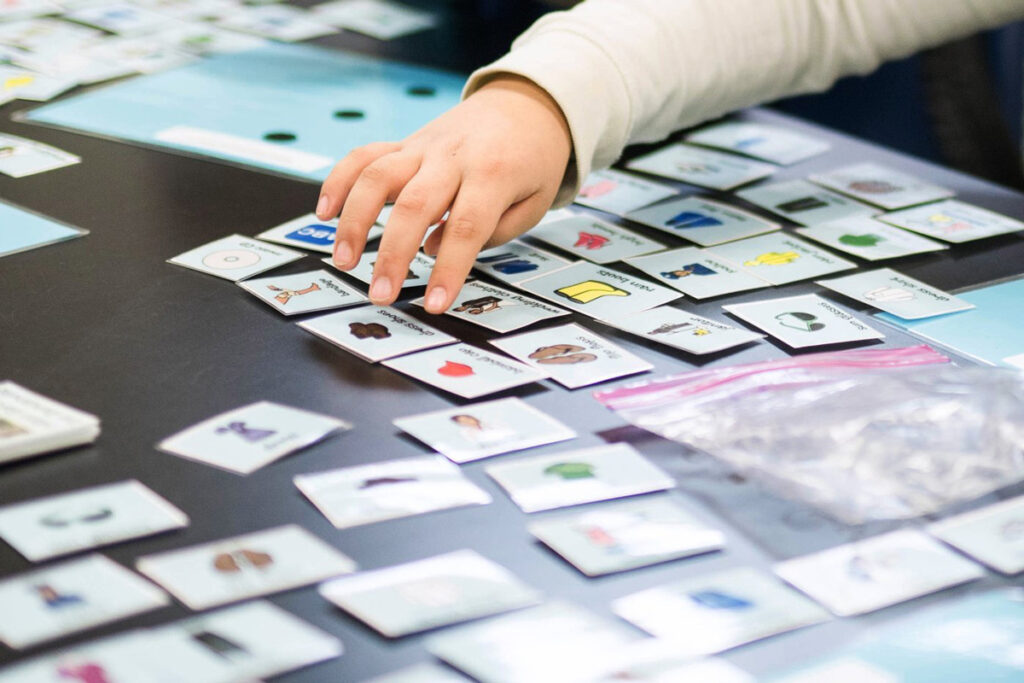 Exemplo de tecnologia assistiva de baixa tecnologia: foto de várias figuras de comunicação alternativa em uma mesa e uma mão infantil está indo pegar uma delas