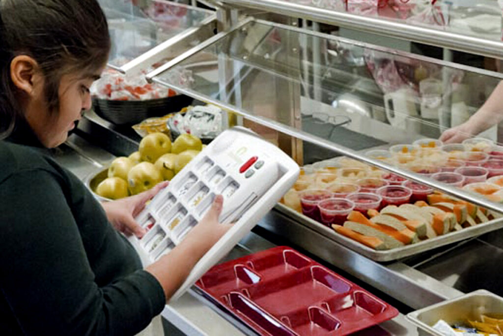 Exemplo de tecnologia assistiva de média tecnologia: foto de uma menina usando seu vocalizador para pedir a comida em uma cantina de escola
