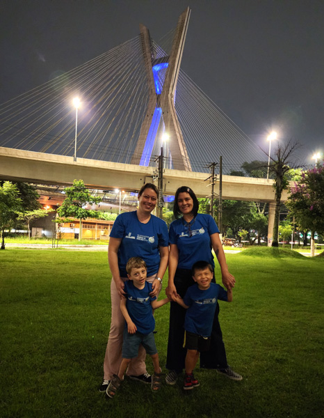 Foto da ponte estaiada iluminada em azul. Em frente a ela posando para a foto estão Fernanda e Priscila com seus filhos vestindo uma camiseta azul
