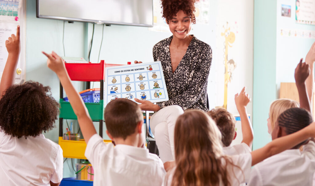 Foto de uma professora mostrando uma prancha de CAA e vários alunos estão levantando a mão para falar.
