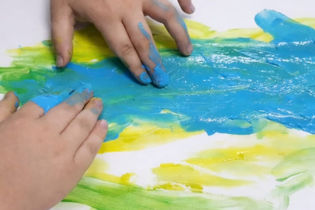 Foto de uma criança fazendo uma pintura nos tons de azul, amarelo e verno com as mãos em um fundo branco.
