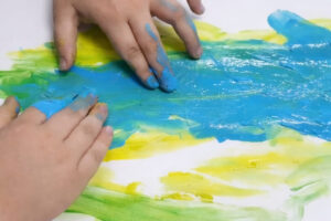 Foto de uma criança fazendo uma pintura nos tons de azul, amarelo e verno com as mãos em um fundo branco.