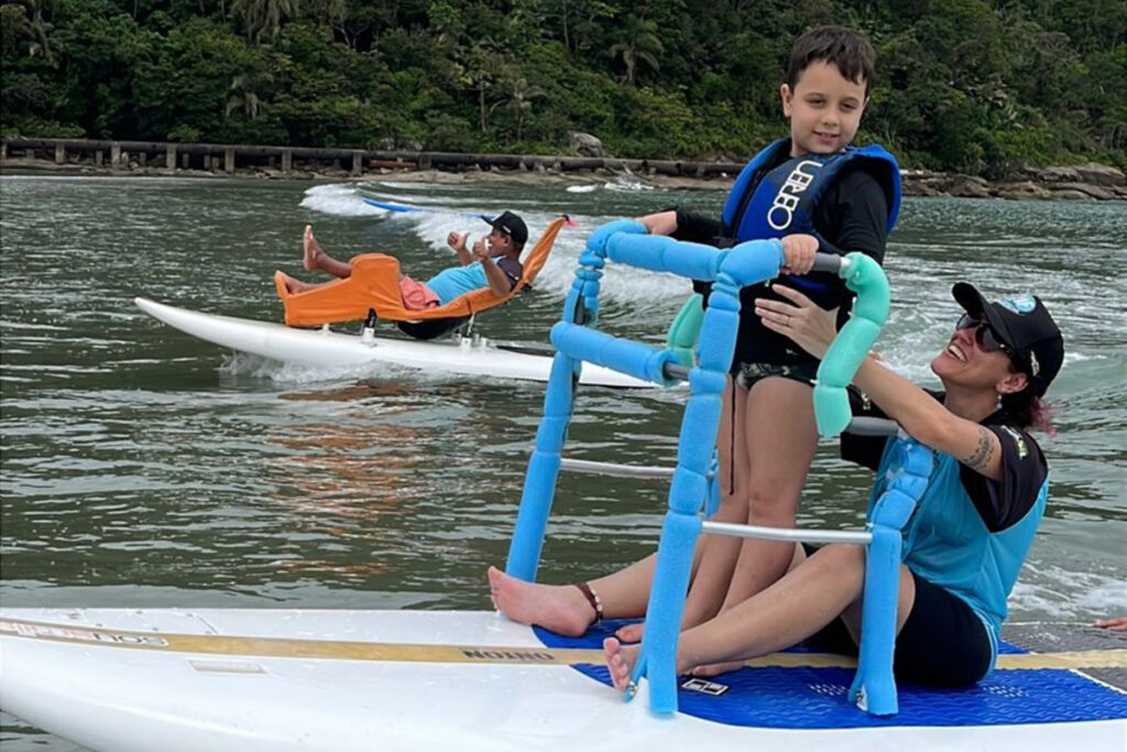 Foto de Dafne segurando uma criança enquanto surfam em uma prancha adaptada.