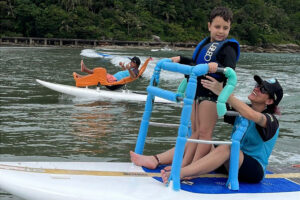 Foto de Dafne segurando uma criança enquanto surfam em uma prancha adaptada.