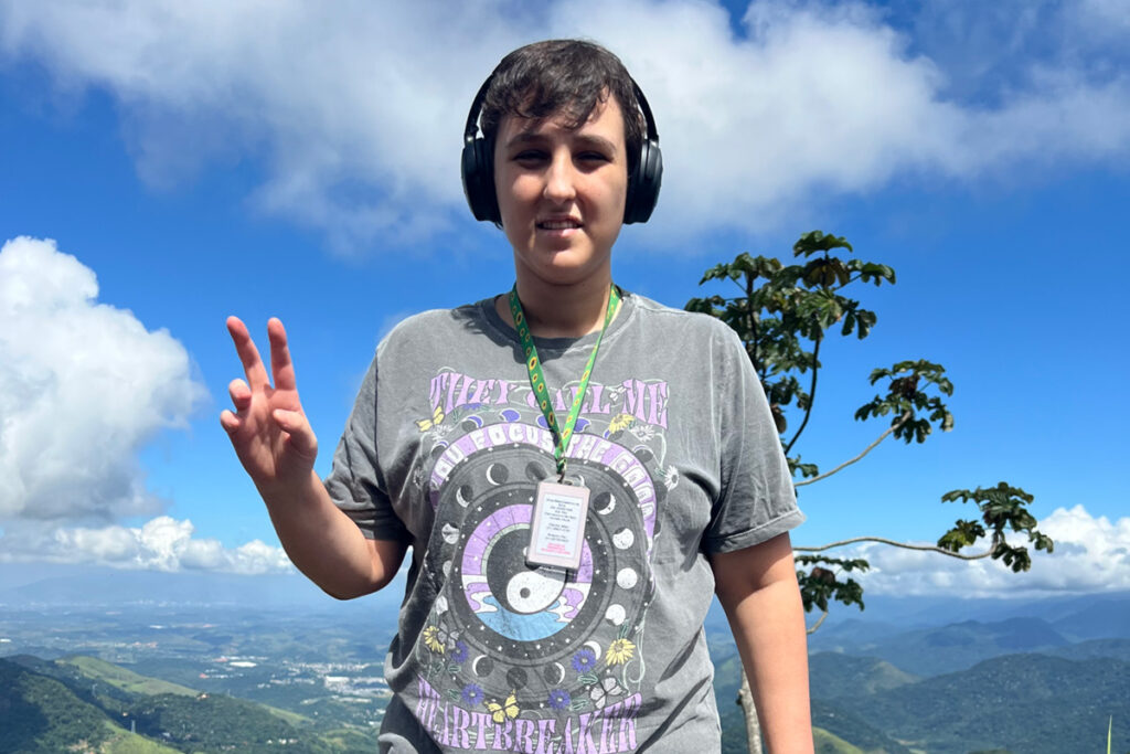 Foto de Alice sorrindo, fazendo o sinal de paz & amor em uma paisagem de um skyline de natureza
