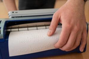 Foto em close de uma máquina de escrever braille com uma mão lendo com os dedos um papel que acabou de ser digitado