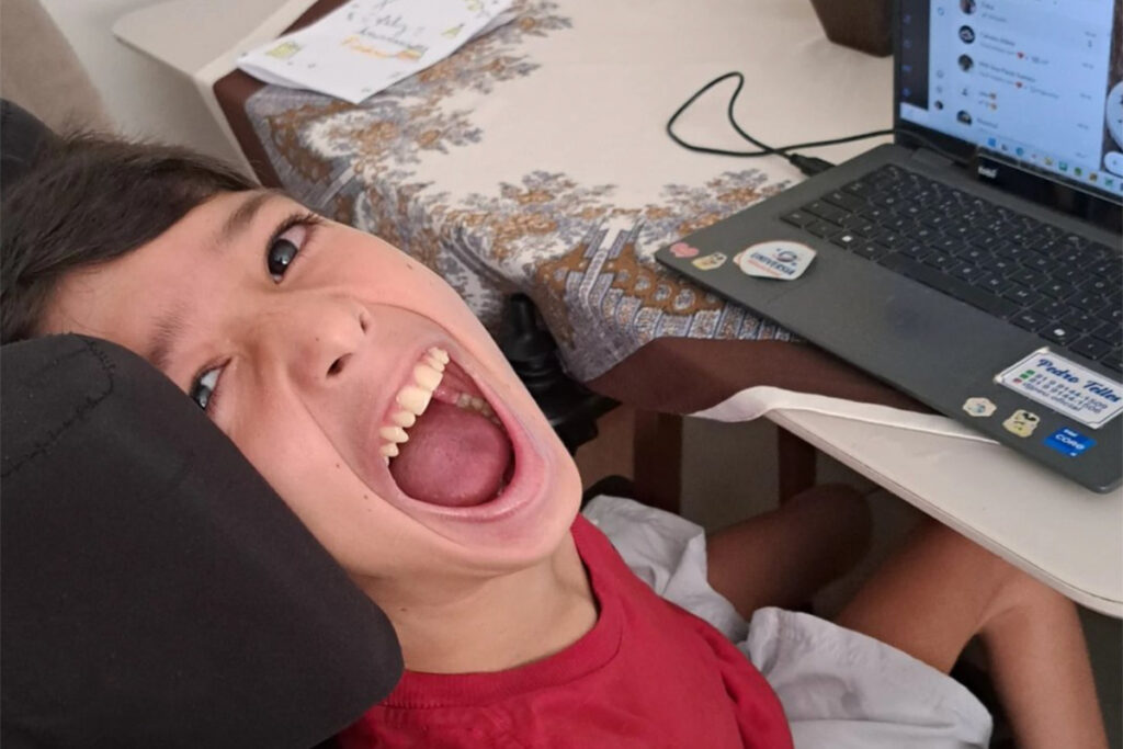 Foto de Pedro sorrindo olhando para a câmera. Eles está sentado em sua cadeira com um tablet com rastreador ocular em sua frente.