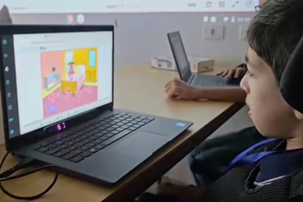 Foto de Pedro na escola em uma mesa ao lado de um colega. Ele está concentrado fazendo um projeto em seu tablet usando o rastreador ocular.