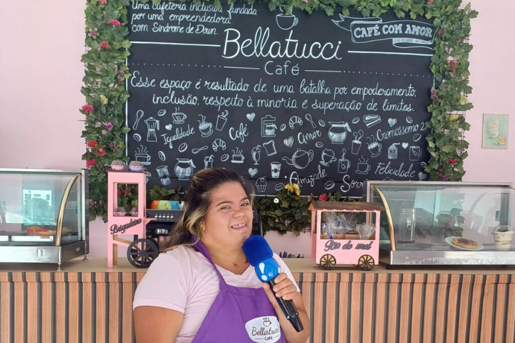 Foto de Jessica com um microfone na mão em frente ao balcão de seu café, com uma lousa com o logo do Bellatucci café cheio de desenhos e palavras sobre inclusão e amor.