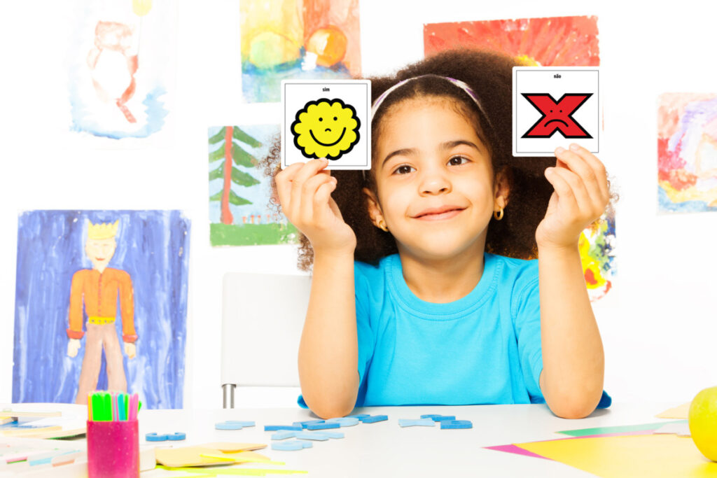 Foto de uma menina em uma mesa cheia de desenhos segurando dois simbolos de CAA: sim e não