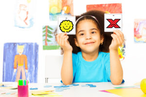 Foto de uma menina em uma mesa cheia de desenhos segurando dois simbolos de CAA: sim e não