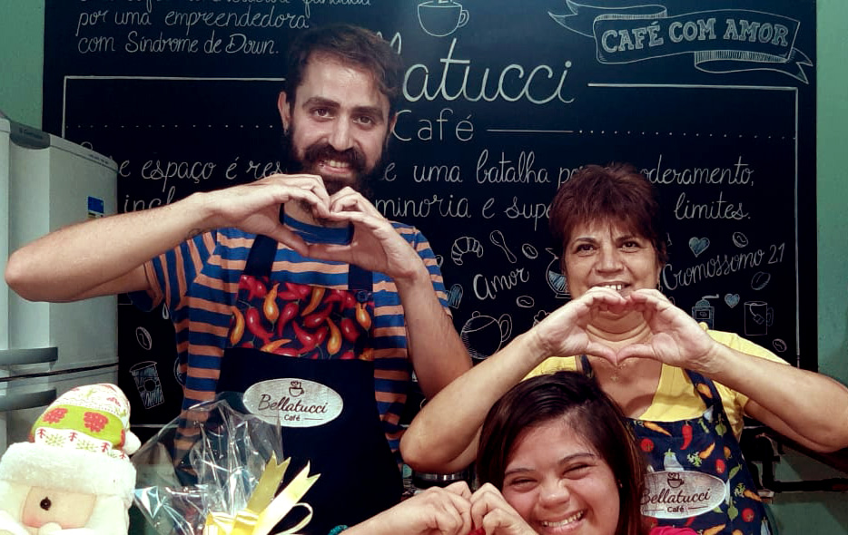 Foto da família com Jessica em frente a uma lousa com logo do Ballatucci Café com desenhos e palavras sobre inclusão e amor. Todos estão fazendo o símbolo do coração com as mãos.