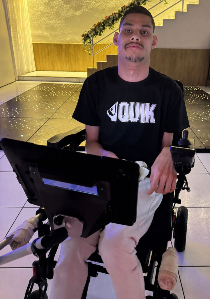 "Matheus Pinheiro sentado em sua cadeira de rodas, utilizando um dispositivo de comunicação com controle ocular acoplado à frente. A foto foi registrada em um ambiente interno iluminado, destacando o uso da tecnologia assistiva para comunicação e autonomia."