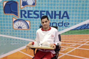 "Matheus Pinheiro em sua cadeira de rodas durante participação em programa esportivo da AACD. Ele está posicionado em frente a um painel temático de esportes, representando sua atuação como atleta da bocha paralímpica."
