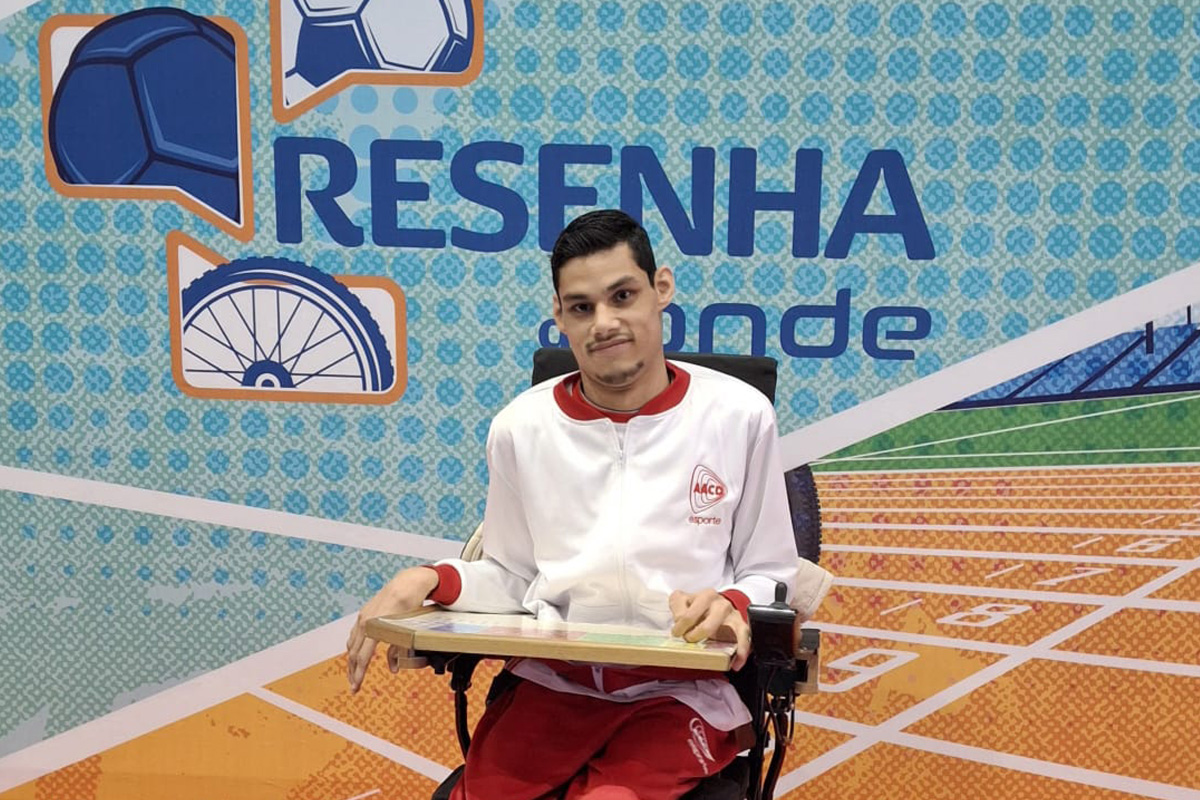 "Matheus Pinheiro em sua cadeira de rodas durante participação em programa esportivo da AACD. Ele está posicionado em frente a um painel temático de esportes, representando sua atuação como atleta da bocha paralímpica."