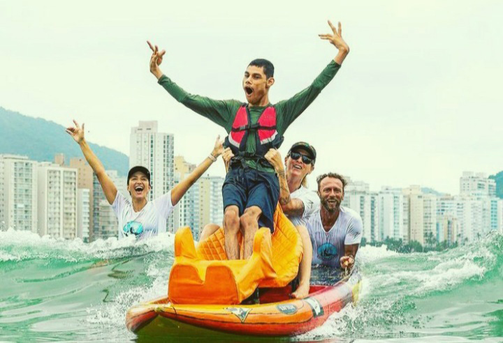 "Matheus Pinheiro participa de uma atividade de surf adaptado no mar, acompanhado por instrutores. Ele aparece sorrindo e comemorando sobre uma prancha adaptada, demonstrando inclusão e participação em esportes radicais."