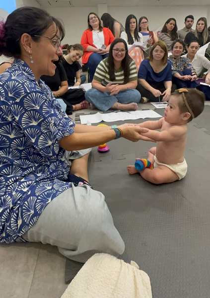 Foto de Dafne brincando com uma bebê bem novinha e várias mulheres estão olhando para aprender
