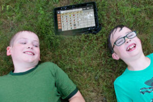 Foto de dois meninos deitados na grama rindo. Acima deles um tablet de Comunicação alternativa todo molhado