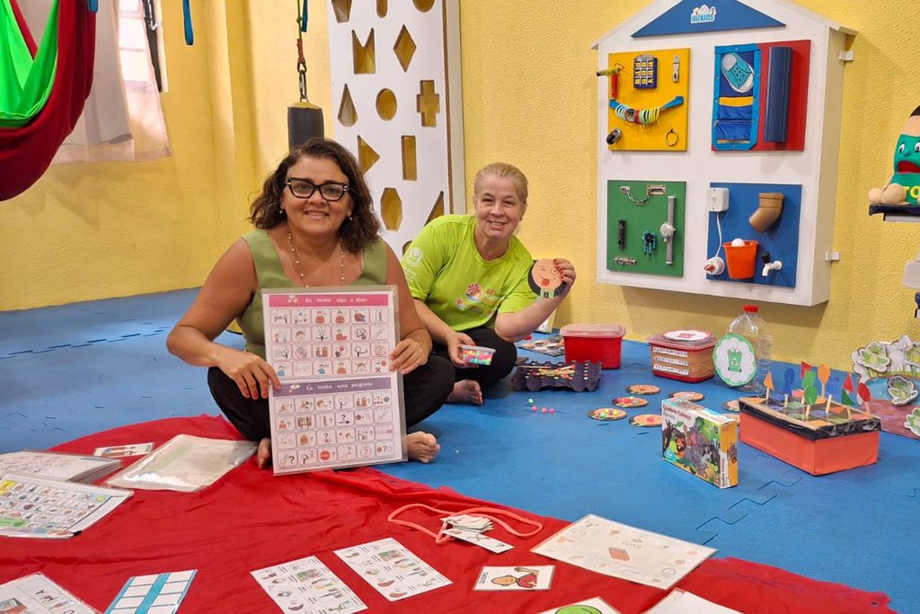 Foto de Adriana e uma outra professora em uma escola com alguns materiais de CAA