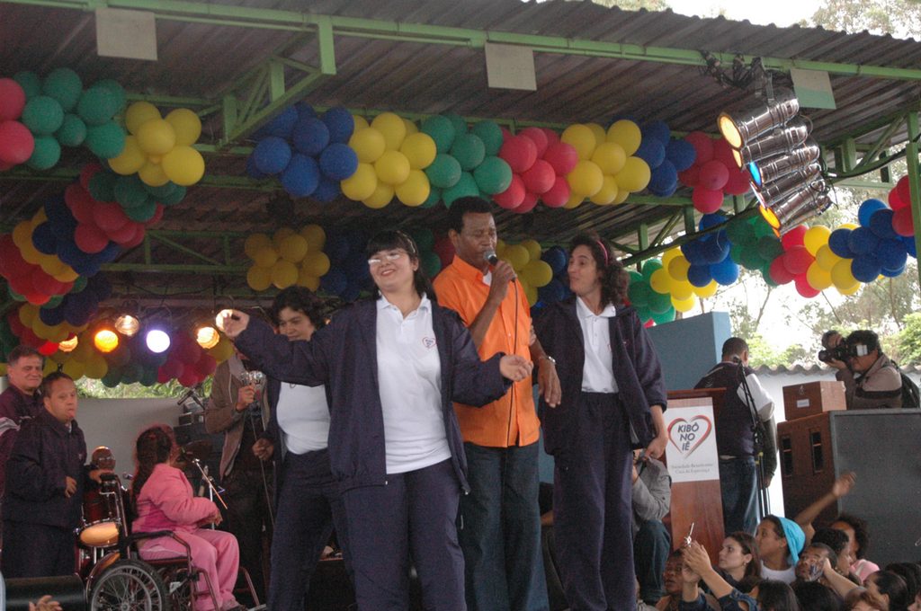 Grupo de pessoas em um palco durante uma apresentação em evento comunitário, com participantes e uma pessoa em cadeira de rodas na plateia.