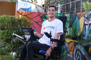 Matheus Pinheiro, atleta paralímpico com paralisia cerebral, sorri sentado em sua cadeira de rodas ao lado de um dispositivo de Comunicação Aumentativa e Alternativa (CAA).