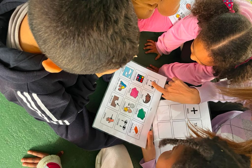Crianças exploram pranchas de comunicação alternativa com símbolos de cores, ações e objetos durante uma atividade coletiva de aprendizagem.