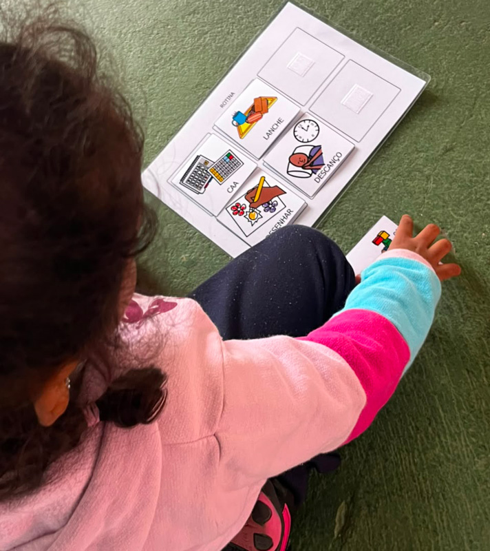 Criança utilizando prancha de comunicação alternativa com símbolos visuais para identificar atividades da rotina escolar, como lanche, descanso e desenhar.